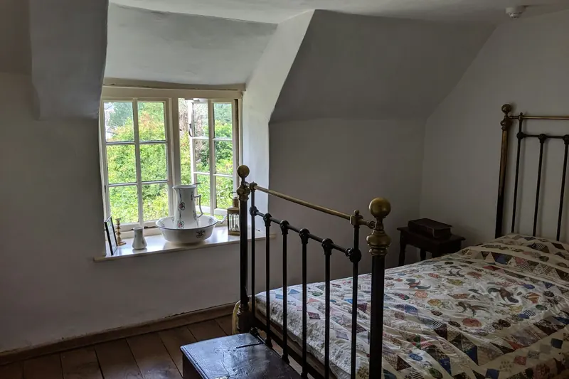 Chambre mansardée avec lucarne, lit en fer forgé couvert d’un dessus-de-lit en patchwork, broc et cuvette en faïence sur le rebord de la fenêtre.