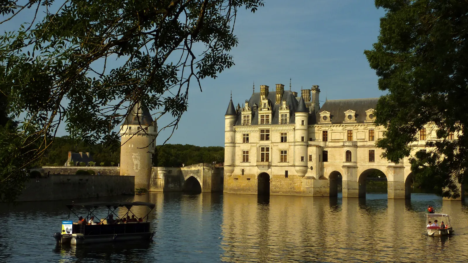 Le château de Chenonceau en fin de journée, vu depuis le Cher, deux bateaux de promenade sur l’eau.