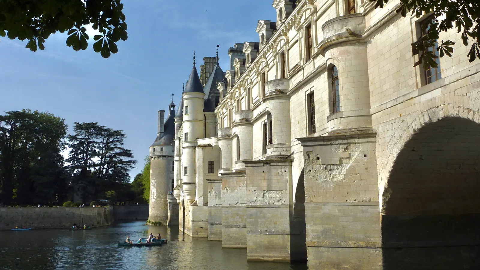 La façade du château de Chenonceau vue d’en bas depuis le Cher, ses arches de pierre et un canoë sur l’eau.