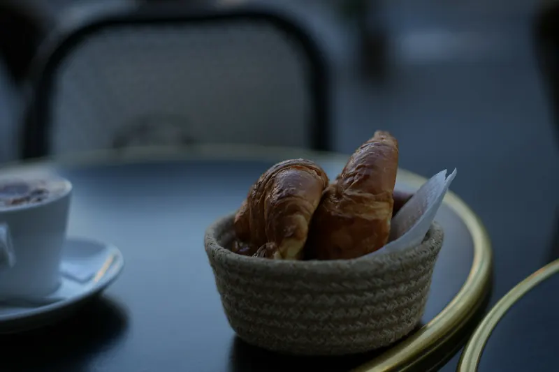 Panier de croissants posé sur une table de bistrot ronde, chaises en fer noir en arrière-plan.